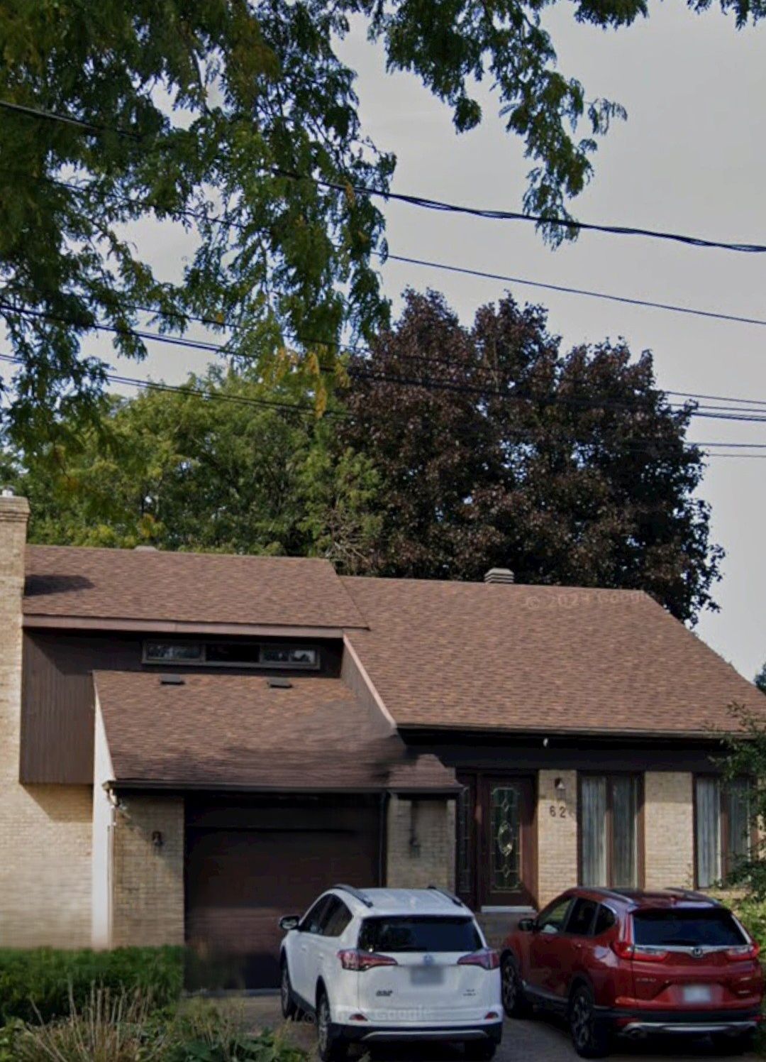 Une maison en briques beiges avec un toit marron et un garage pour une seule voiture, avec un SUV blanc et un SUV rouge garés dans l'allée.