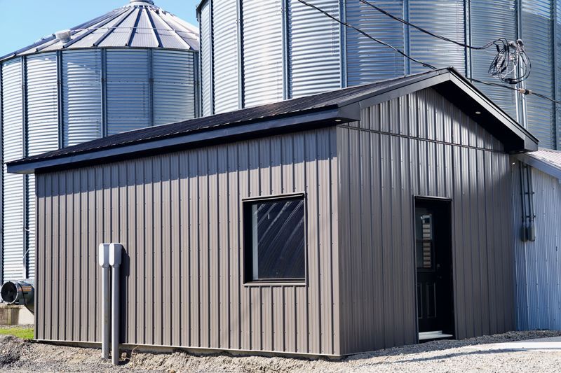 Une petite structure brune aux parois verticales en métal, avec une porte et une fenêtre, placée devant de grands silos à grains argentés.