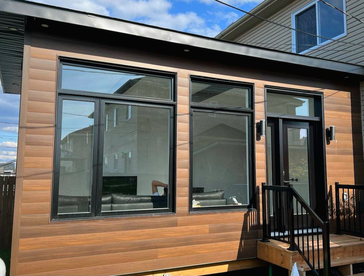 Extérieur de maison moderne avec bardage imitation bois, grandes fenêtres à cadre noir et petite terrasse avec escalier.