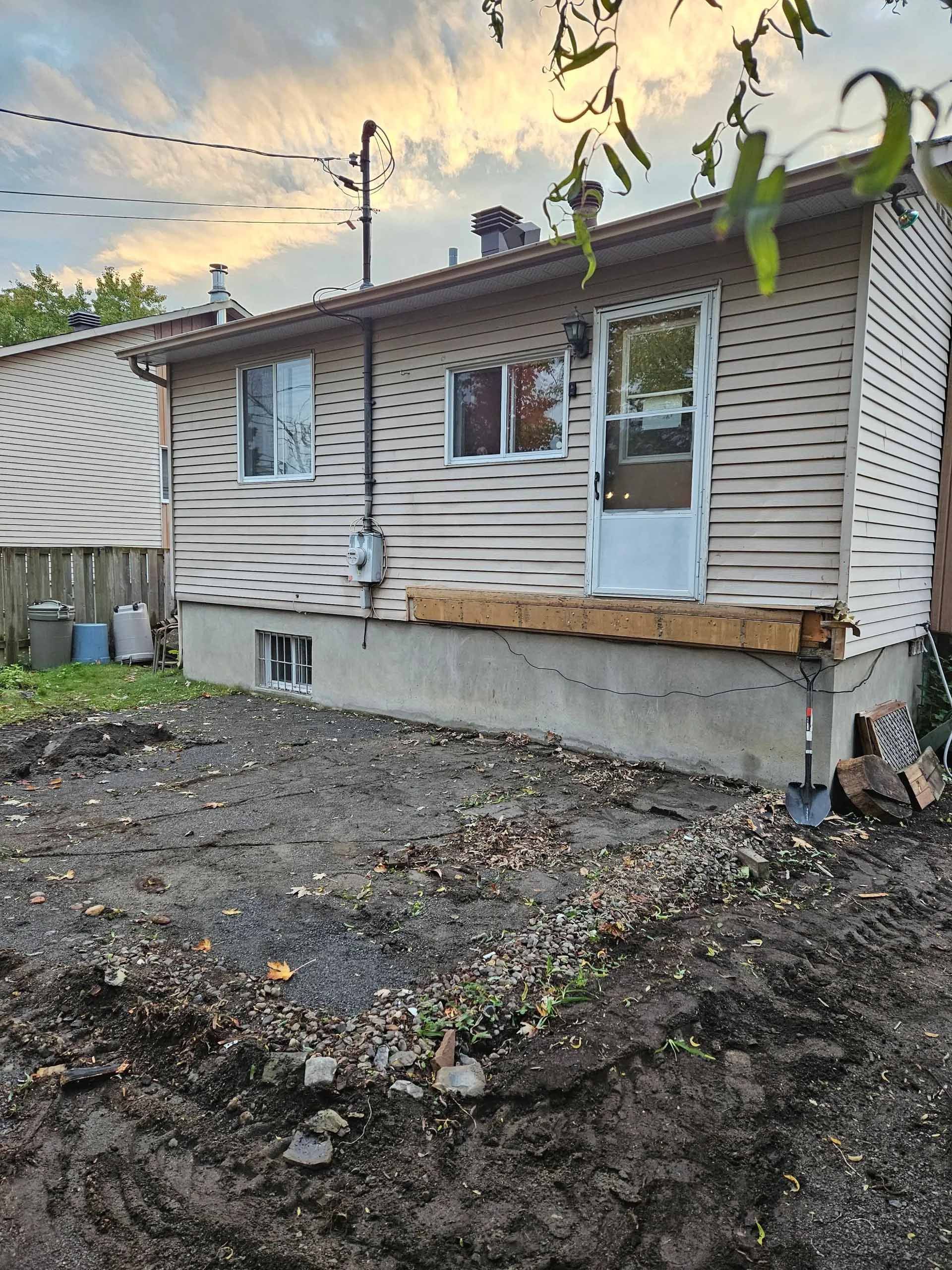 Arrière d'une maison claire avec un jardin nu, une structure de terrasse en bois apparente et une porte moustiquaire blanche sous un ciel de coucher de soleil.