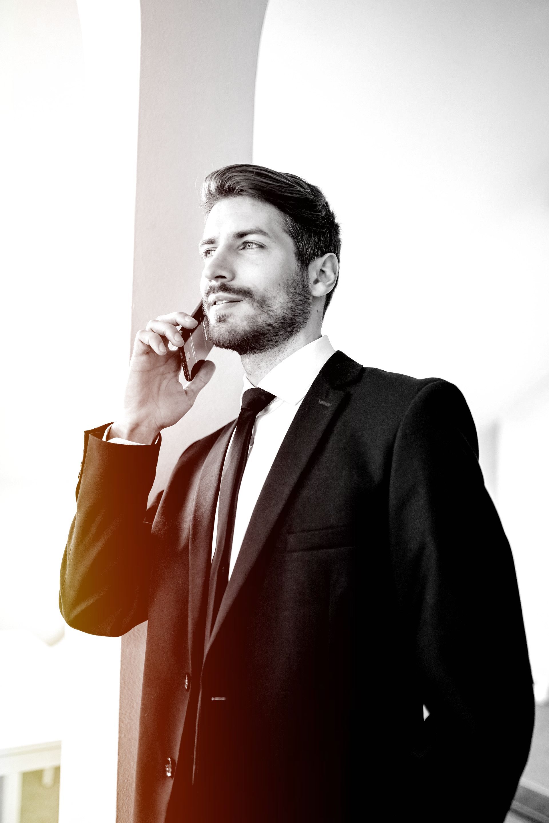 Man in a black suit speaking on a phone in a bright office setting