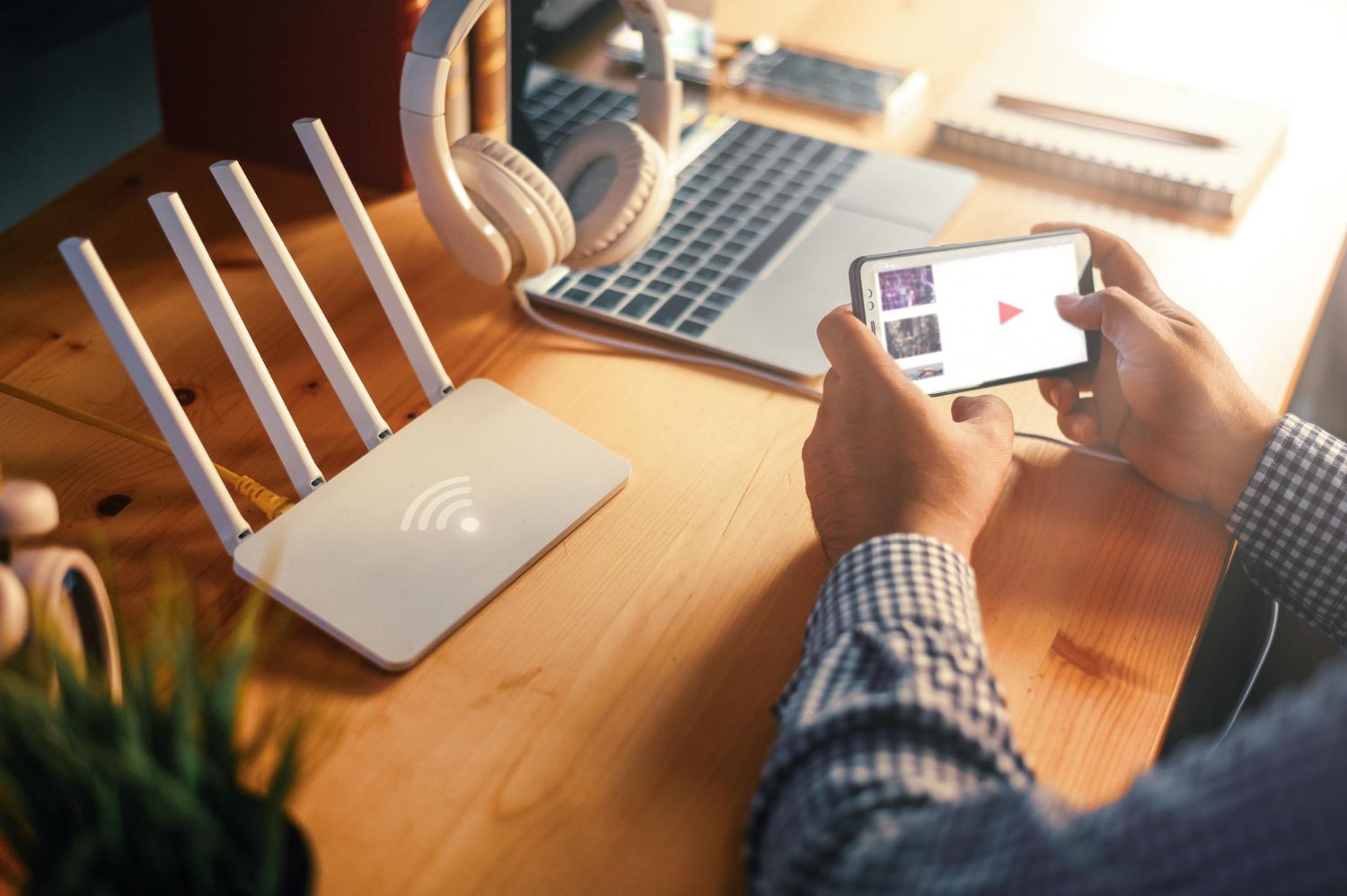 Person using a smartphone beside a Wi‑Fi router and laptop on a wooden desk