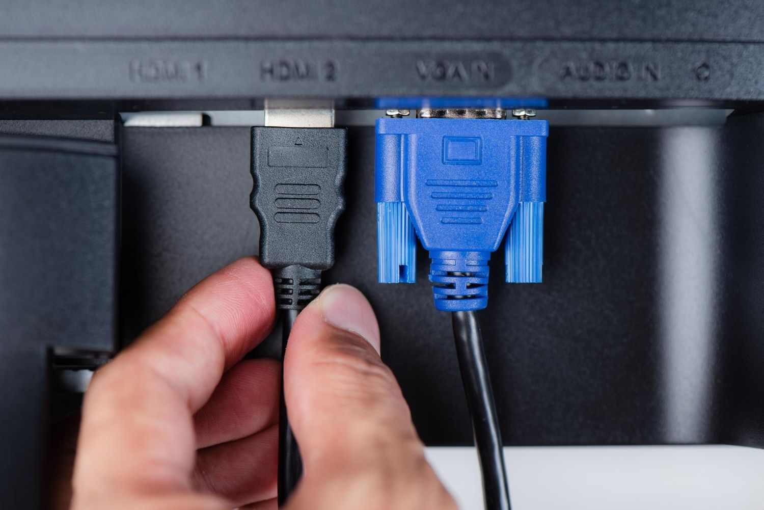 Hand plugging a black cable into an HDMI port beside a blue VGA cable on a monitor