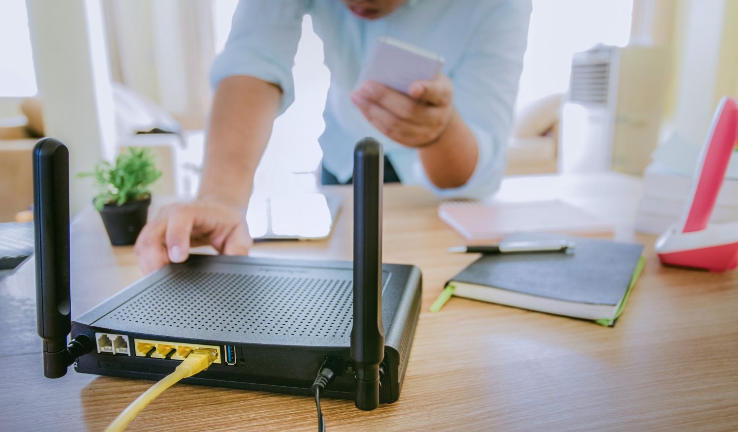 Hands adjusting a black Wi‑Fi router with antennas on a desk, with a laptop and notebooks nearby.