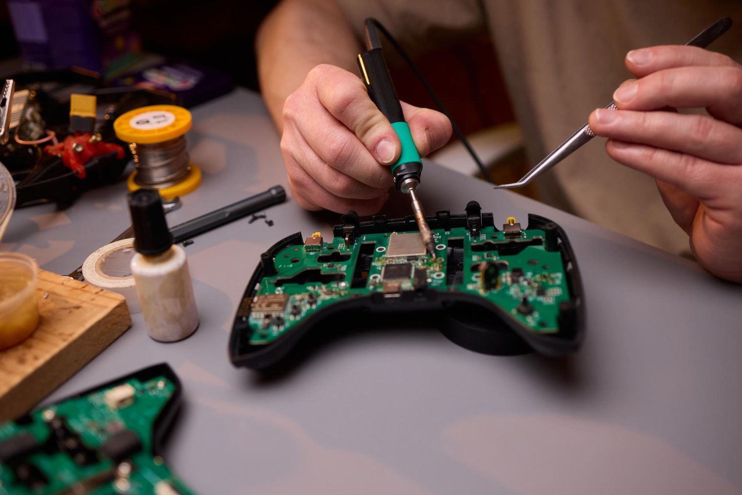 Hands repairing a black game controller with a soldering iron on a workbench