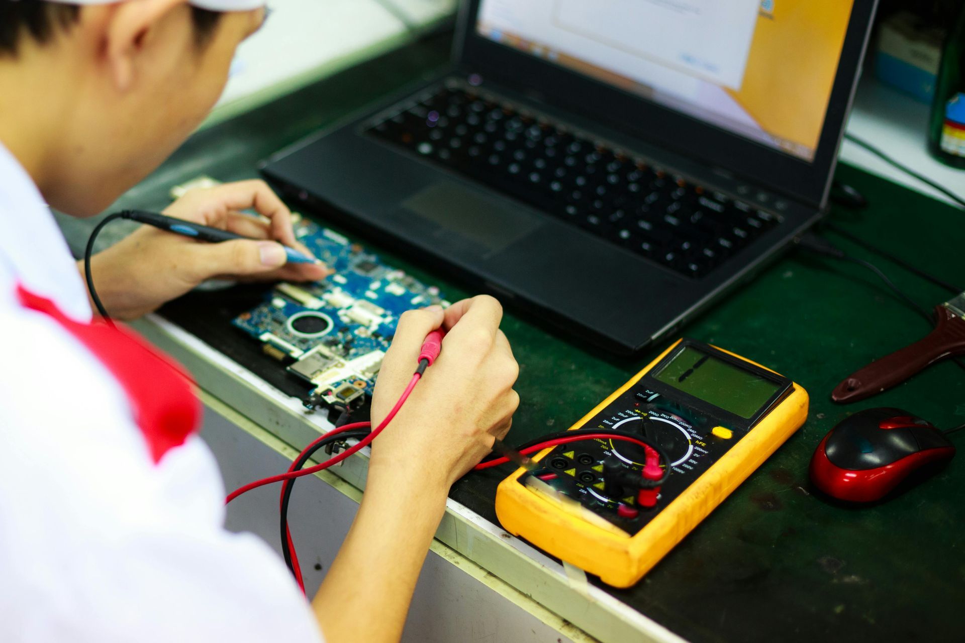 Technician testing a circuit board with a multimeter beside a laptop on a workbench