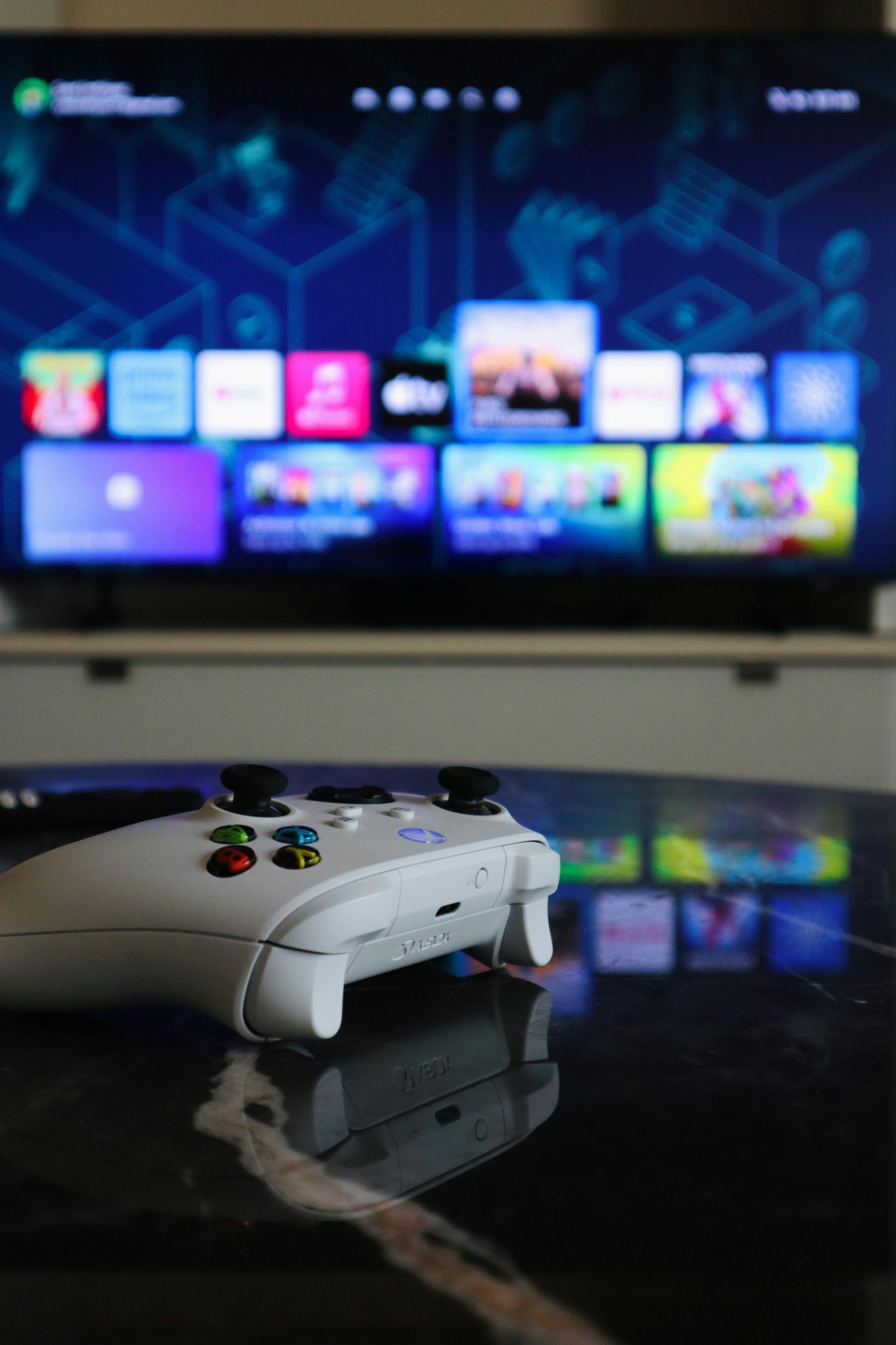 White game controller on a glossy table in front of a blurred TV showing a colorful gaming menu.