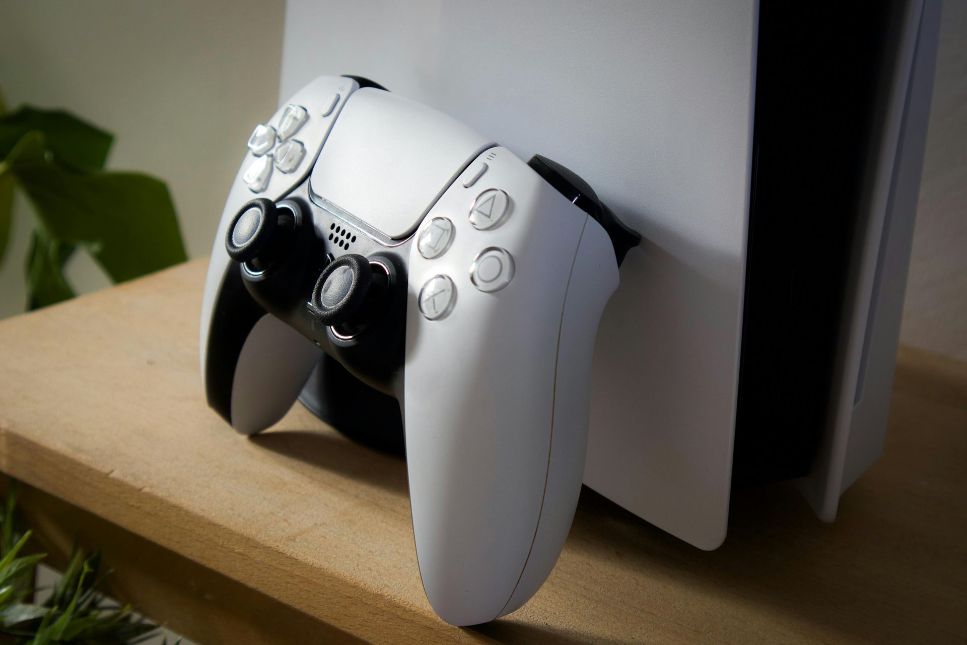 White PlayStation 5 DualSense controller resting against a white console on a wooden shelf