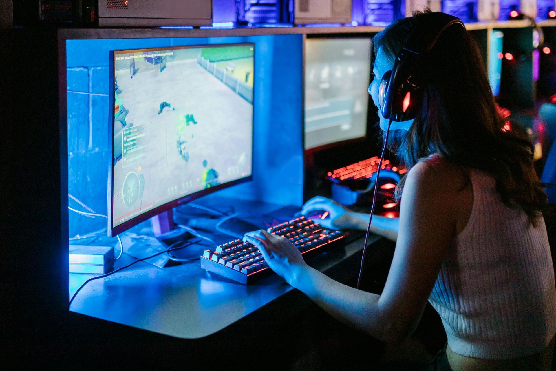 Person gaming at a desktop PC in a neon-lit room, viewed from behind, with a bright blue monitor and keyboard.
