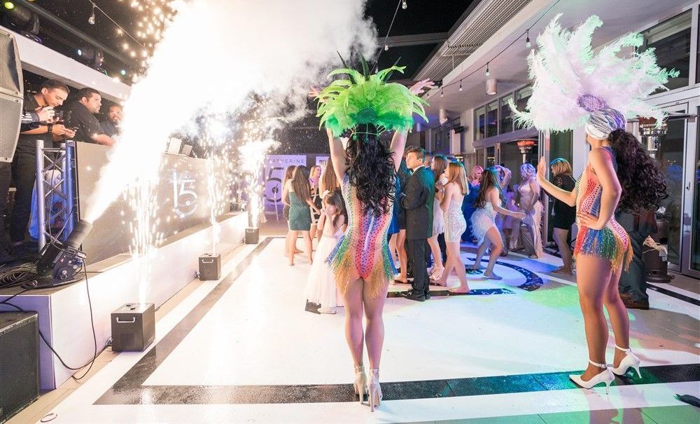 Dancers in bright feathered costumes perform on a lit nightclub stage with a crowd nearby