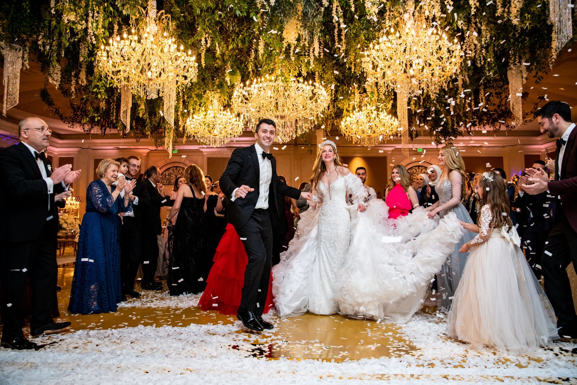Newlyweds dance under chandeliers as guests cheer and applaud in an elegant ballroom.