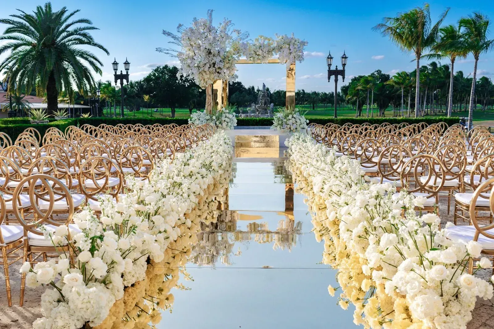 Outdoor wedding aisle with white flowers, gold chairs, and reflecting water under palm trees