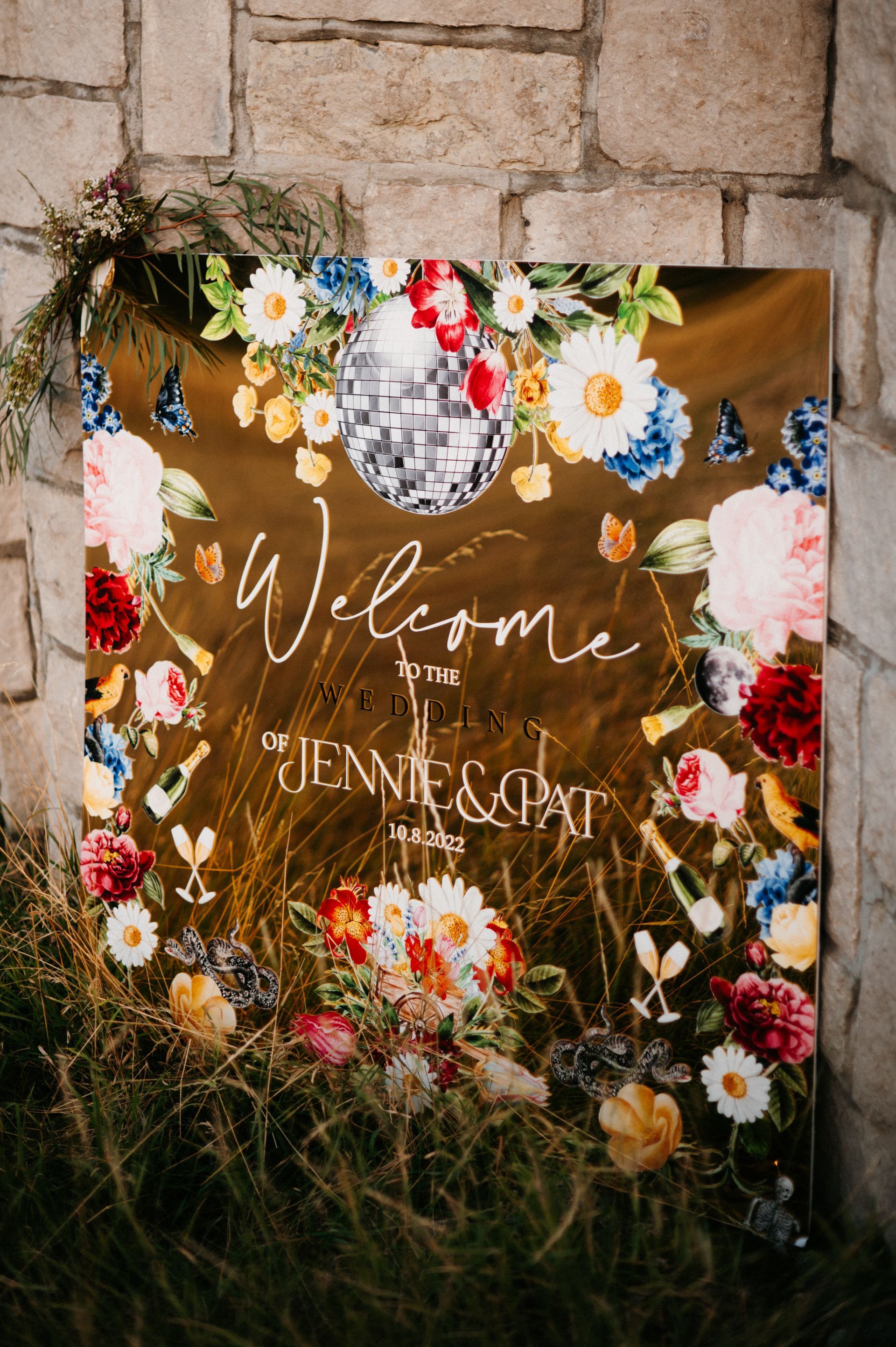 Floral welcome sign on a stone wall, surrounded by grass and colorful flowers, reading “Welcome” and names below