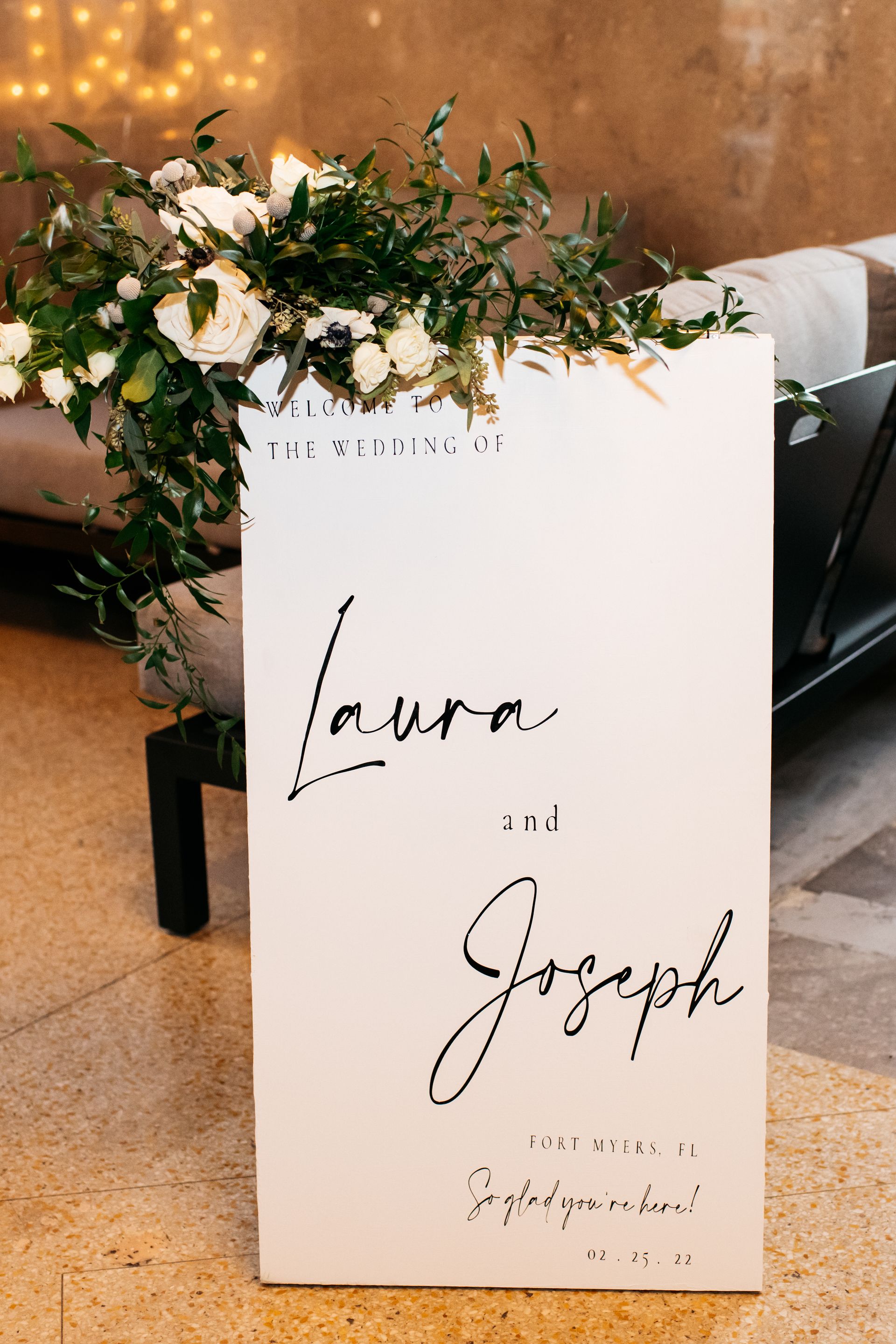 White wedding sign with elegant script, surrounded by green and white floral arrangement.
