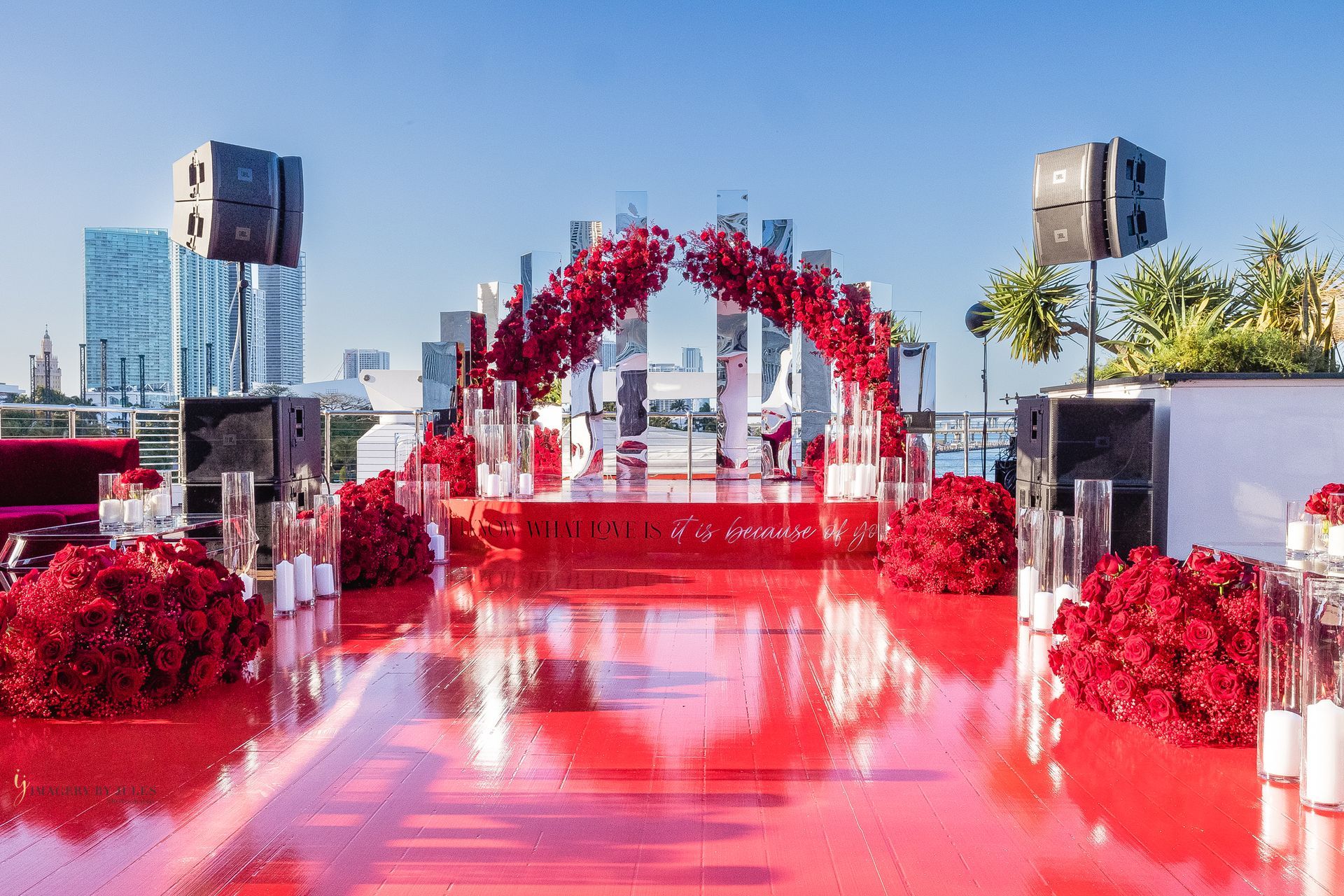 Red carpet walkway with a red floral arch and speakers at an outdoor event under a clear blue sky