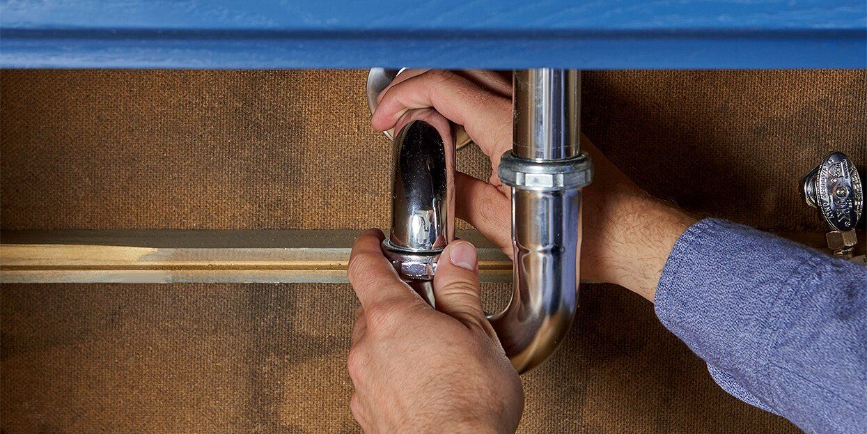A pair of hands adjusts a metallic plumbing pipe under a sink against a textured, light-brown wall.