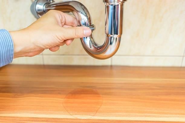 A hand touches a chrome P-trap pipe dripping onto a wooden surface, indicating a leak beneath a sink.
