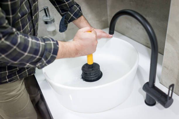 A person uses a yellow-handled black plunger to clear a drain in a white bathroom sink with a black faucet.