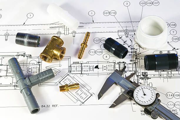 A person in a red shirt holding a bucket under a leaking water heater mounted on a tiled wall. Various plumbing fittings, connectors, and a dial caliper arranged on top of a technical engineering blueprint.
