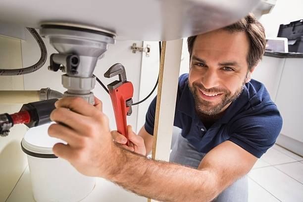A plumber smiles while using a red pipe wrench to fix the plumbing beneath a sink.