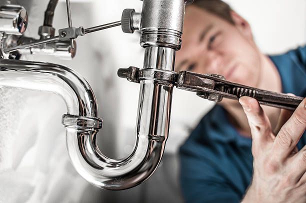A person in a blue shirt uses a wrench to tighten a charome pipe connection under a sink. A person in a blue shirt uses a wrench to tighten a charome pipe connection under a sink.