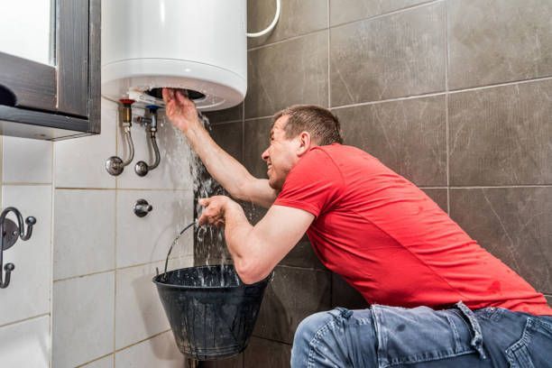 A person in a red shirt holding a bucket under a leaking water heater mounted on a tiled wall. A person in a red shirt holding a bucket under a leaking water heater mounted on a tiled wall.