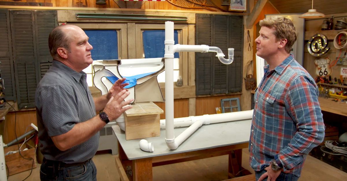 Two people in a workshop discuss a plumbing display featuring white PVC pipes and a clear P-trap on a wooden table. Two people in a workshop discuss a plumbing display featuring white PVC pipes and a clear P-trap on a wooden table.