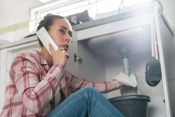 A person talks on a phone while using a cloth to wipe underneath a sink into a bucket. A person talks on a phone while using a cloth to wipe underneath a sink into a bucket.