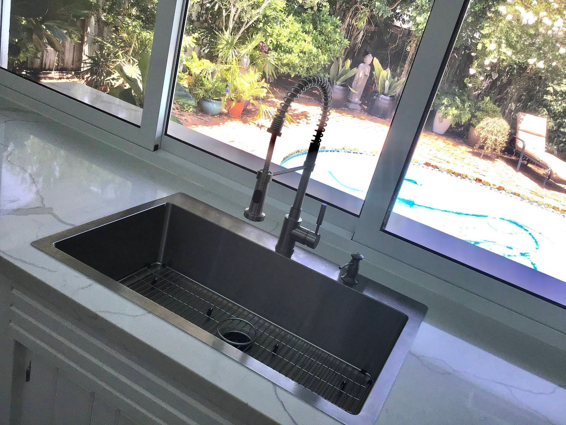 A stainless steel kitchen sink with a tall gooseneck faucet, set into a light-colored countertop before a large window. A stainless steel kitchen sink with a tall gooseneck faucet, set into a light-colored countertop before a large window.