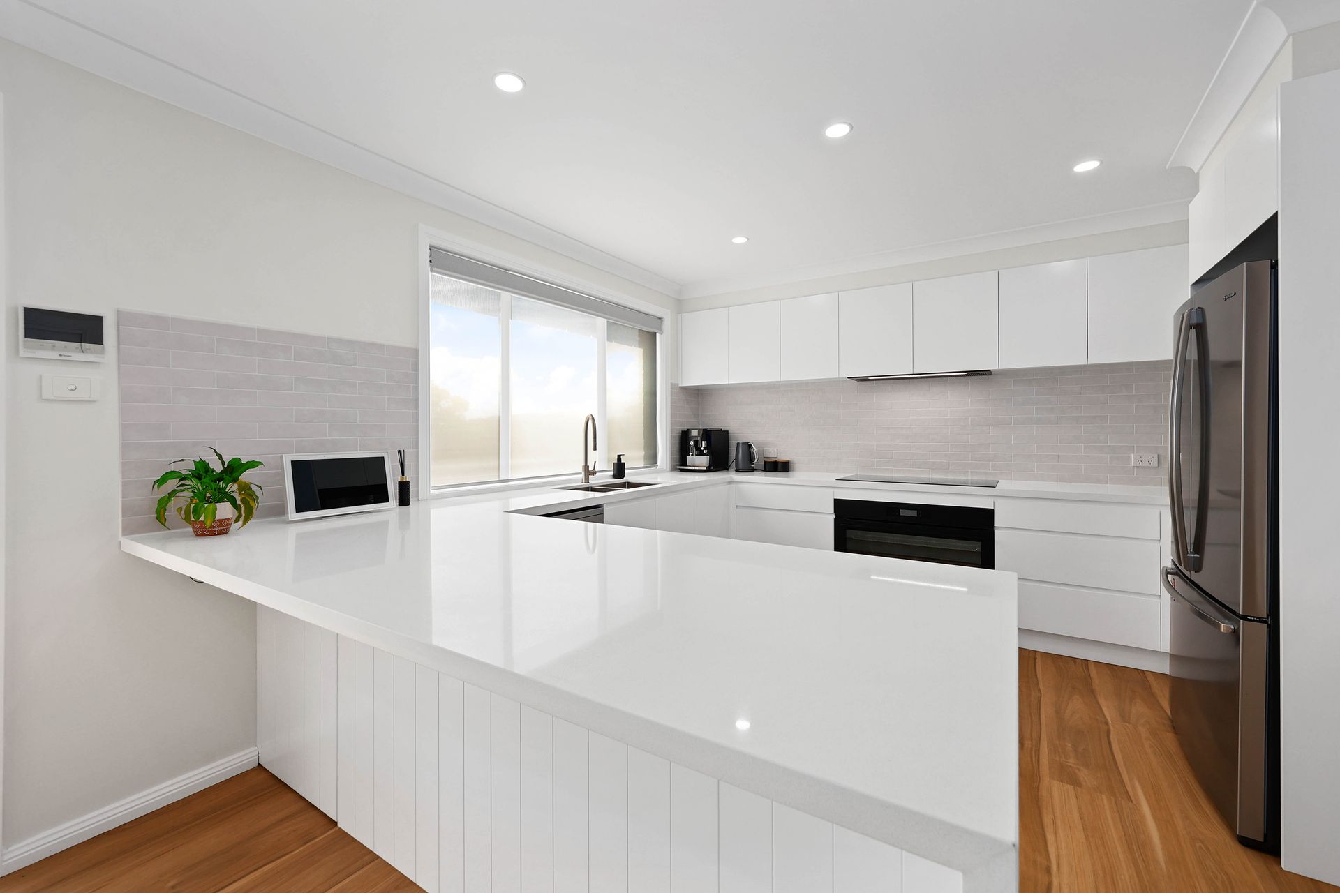 Modern white kitchen with a large island, stainless steel refrigerator, and wooden floor — Above & Beyond Interiors in Mount Annan, NSW