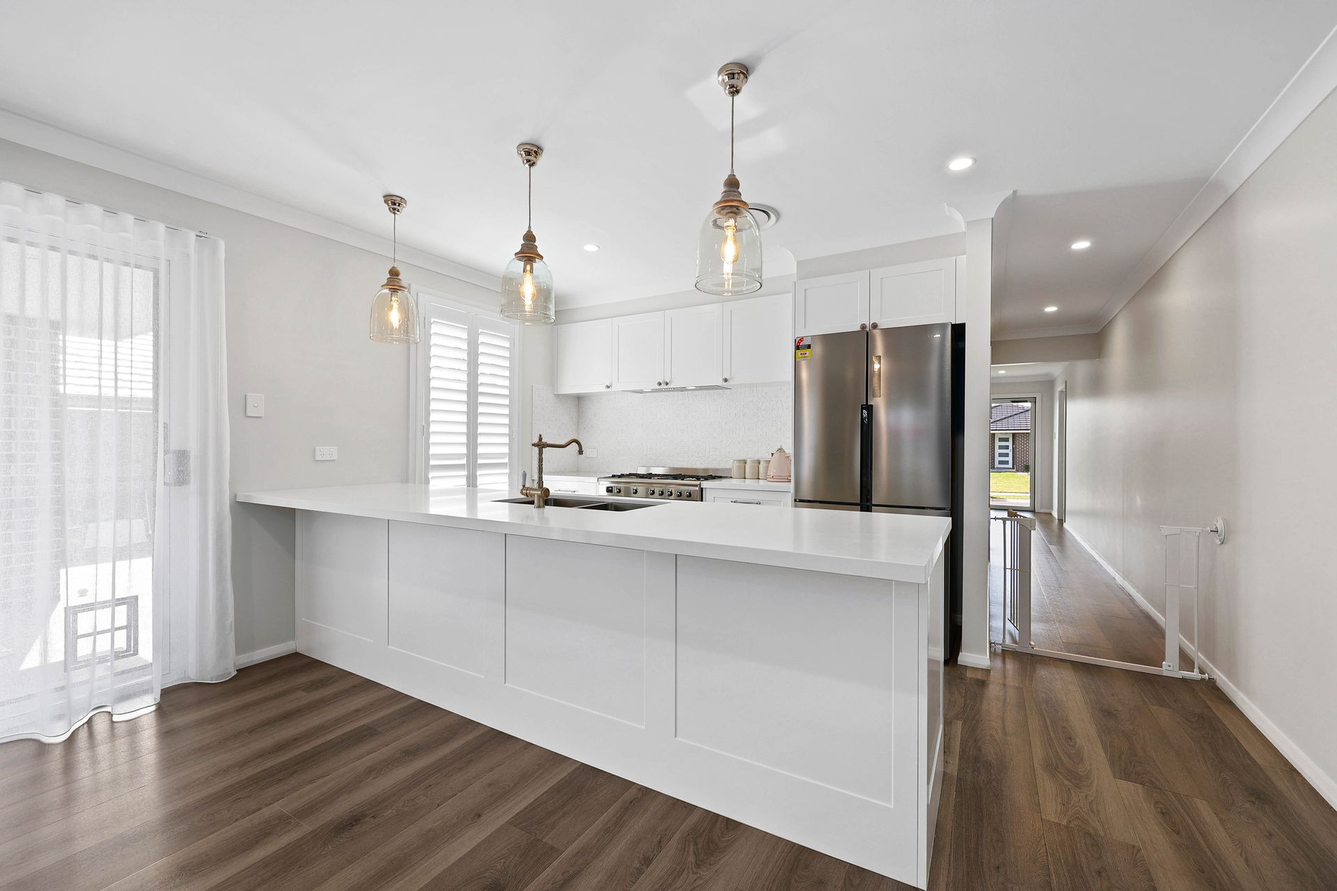 Modern white kitchen with island, pendant lights, and stainless steel fridge; wood floors — Above & Beyond Interiors in Picton, NSW