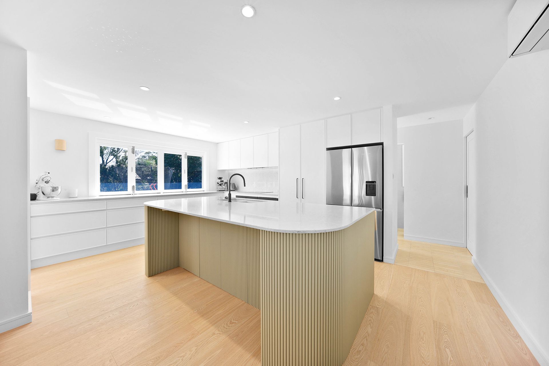 Bright white kitchen with a light tan, ribbed island and stainless steel fridge — Above & Beyond Interiors in Smeaton Grange, NSW