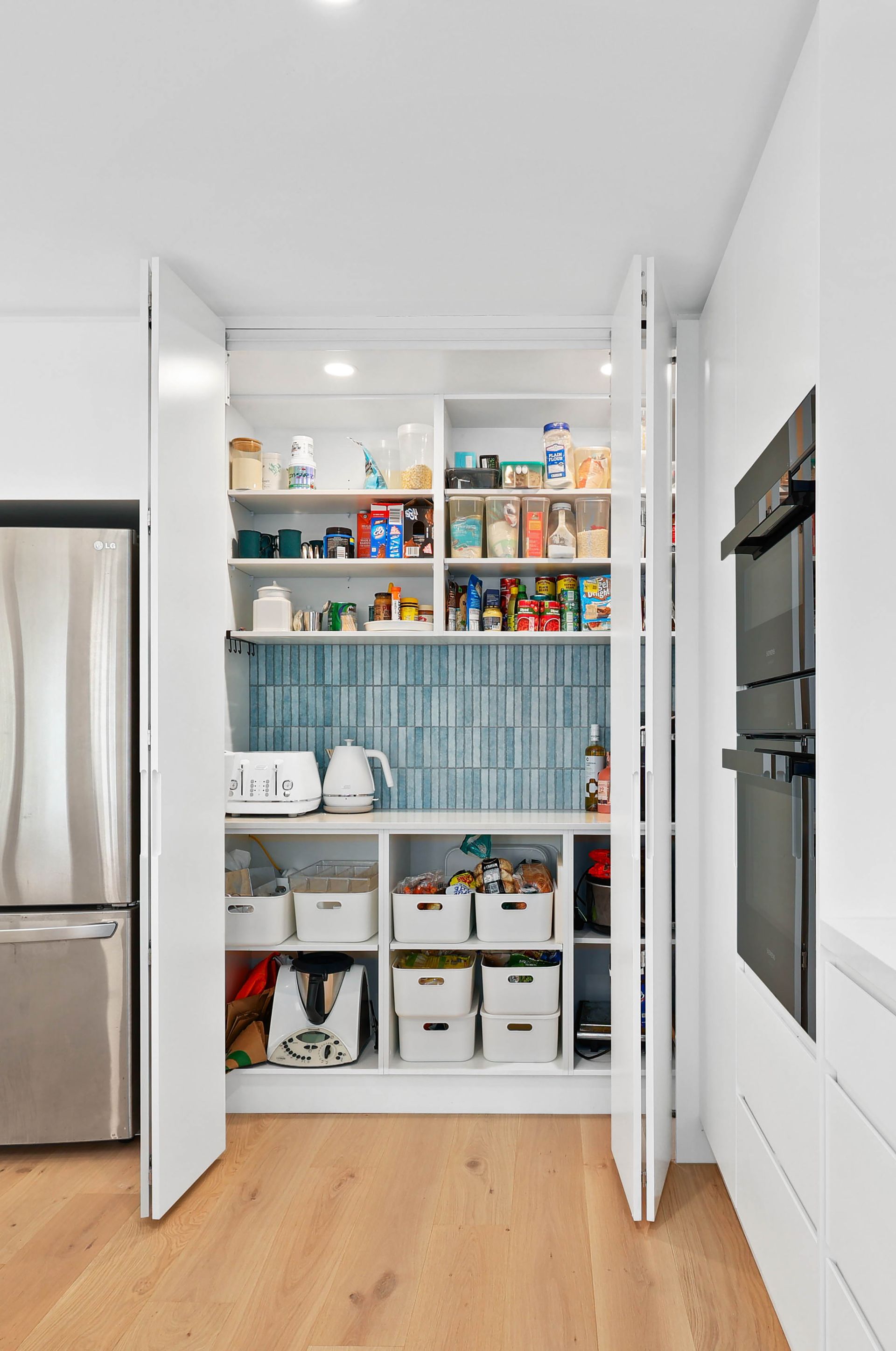 White pantry with open doors, filled with organized food and appliances; next to a fridge and built-in oven — Above & Beyond Interiors in Mount Annan, NSW