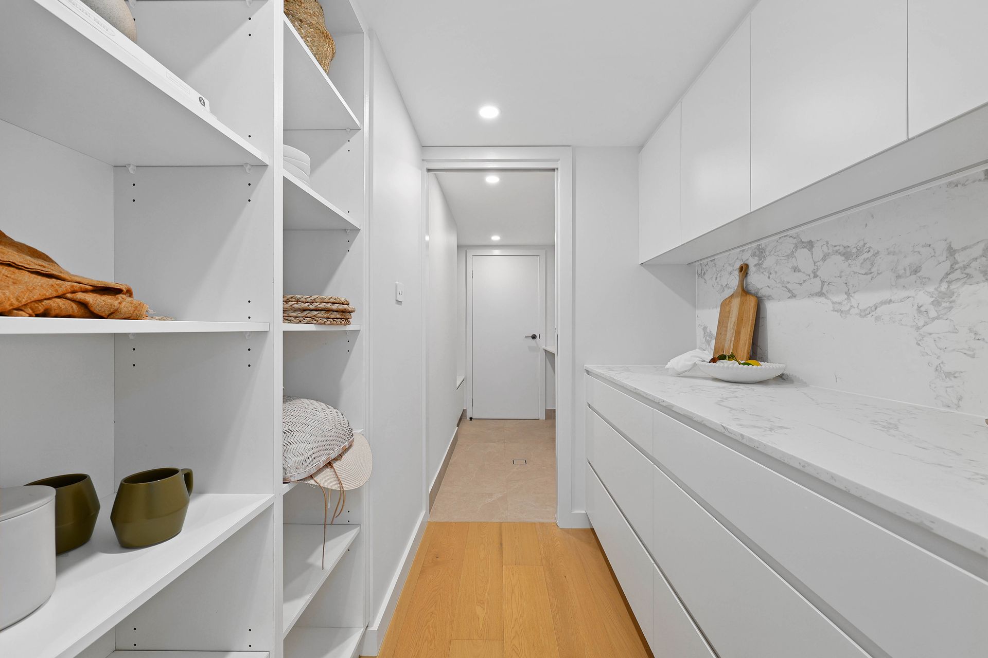 White pantry with shelves on one side, cabinets and countertop on the other, and a doorway at the end — Above & Beyond Interiors in Southern Highlands, NSW