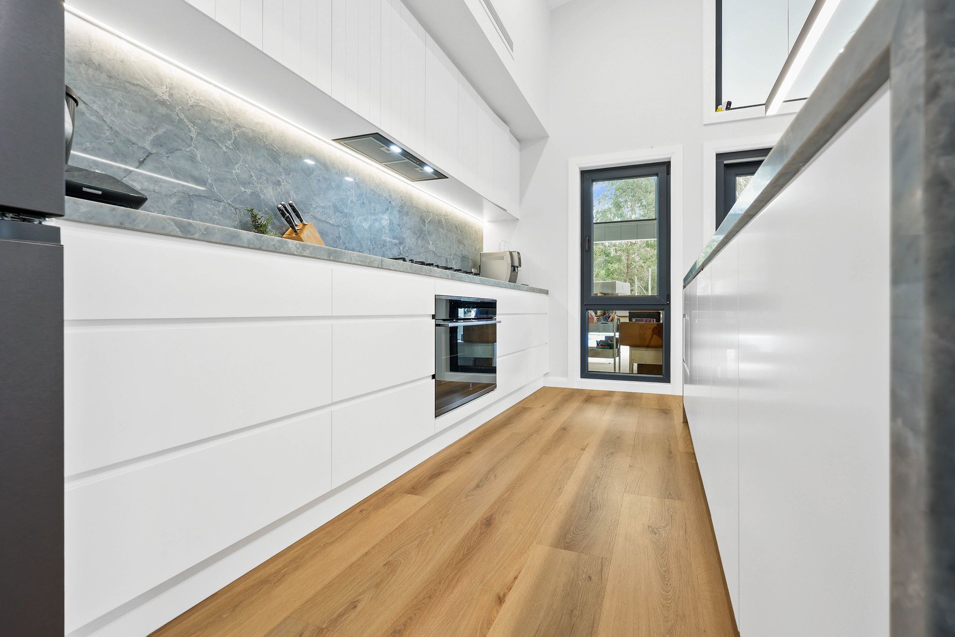 Modern white kitchen with wood floors, stainless steel appliances, and a view of the outdoors — Above & Beyond Interiors in Southern Highlands, NSW