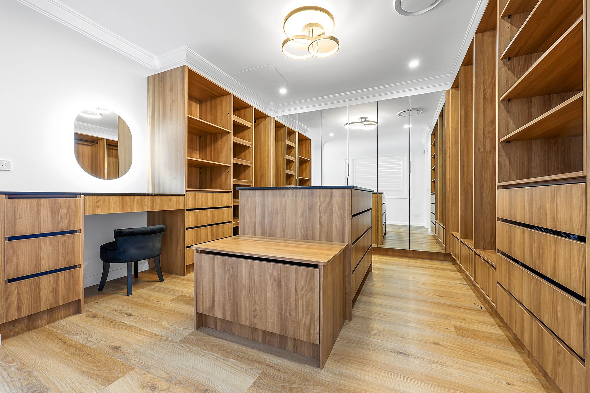 Walk-in closet with wood shelves, drawers, and a vanity. Neutral tones, sleek design, and a central island — Above & Beyond Interiors in Smeaton Grange, NSW