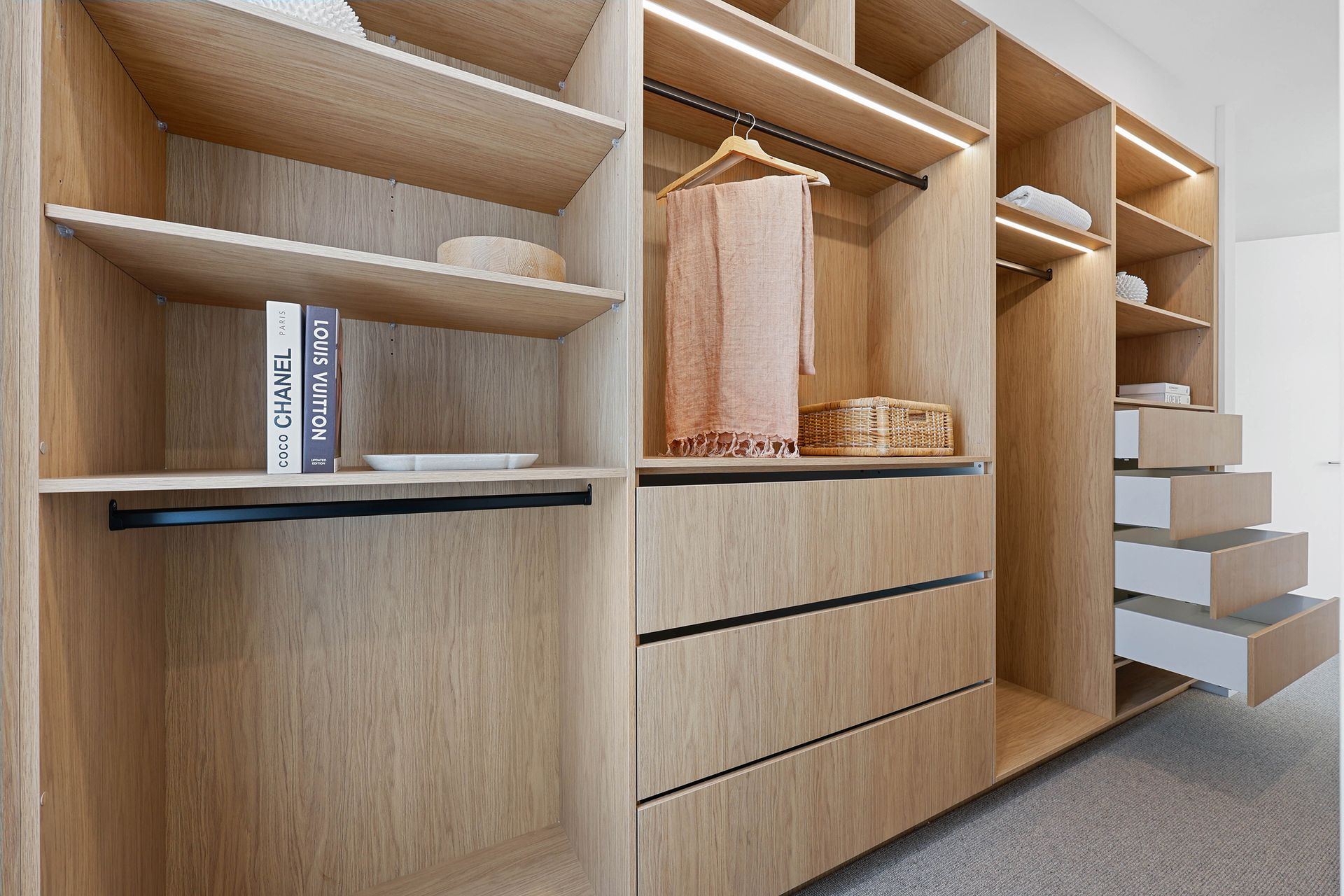 Wooden walk-in closet with open shelves, drawers, and hanging rod. Contains clothes, books, and decorative baskets.