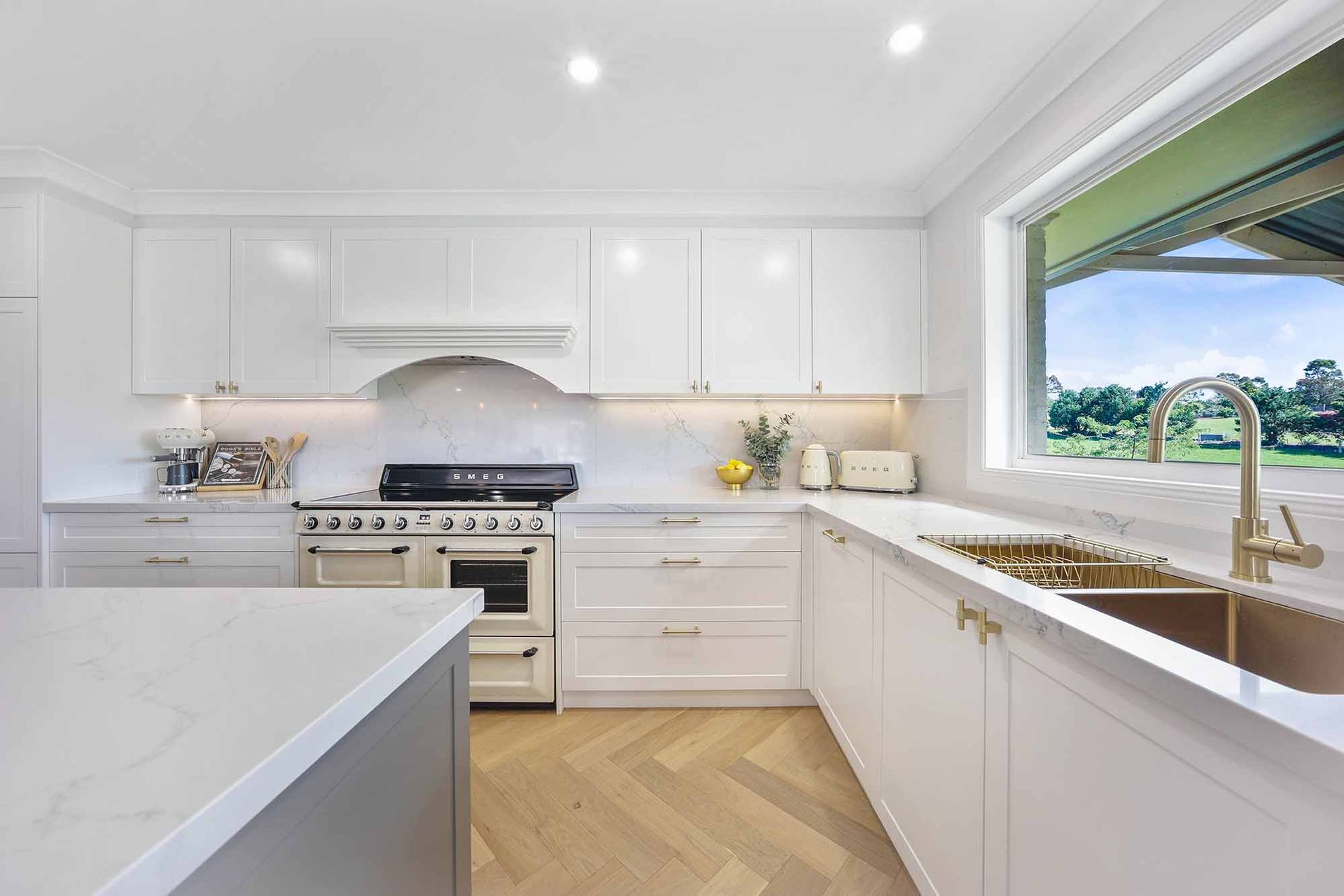 White kitchen with marble countertops, a window overlooking greenery, and wooden floors — Above & Beyond Interiors in Smeaton Grange, NSW