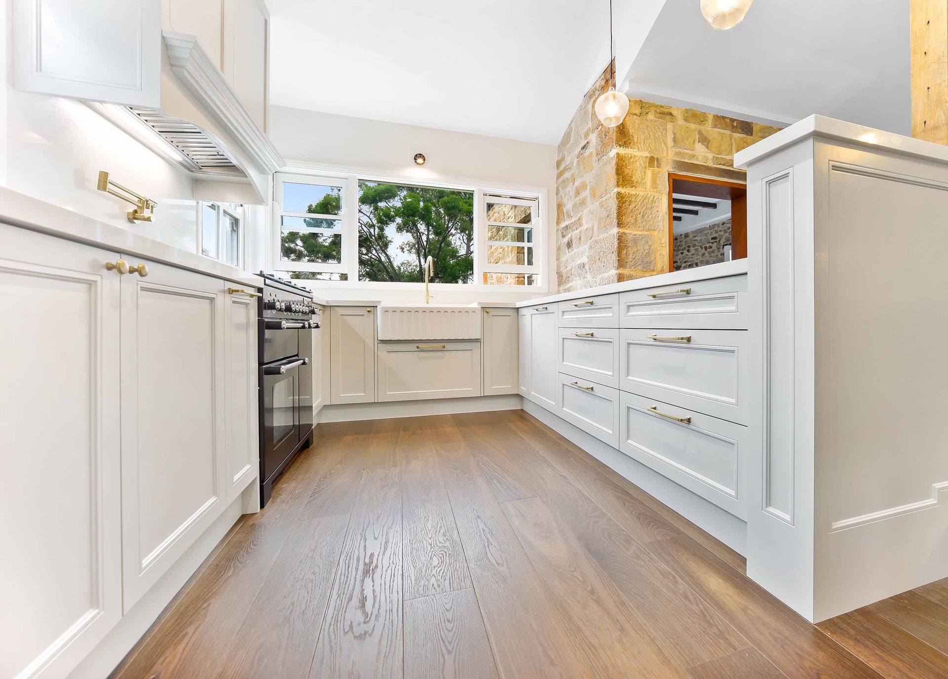 White kitchen with wood floor, a window, stone wall, and white cabinetry — Above & Beyond Interiors in Smeaton Grange, NSW