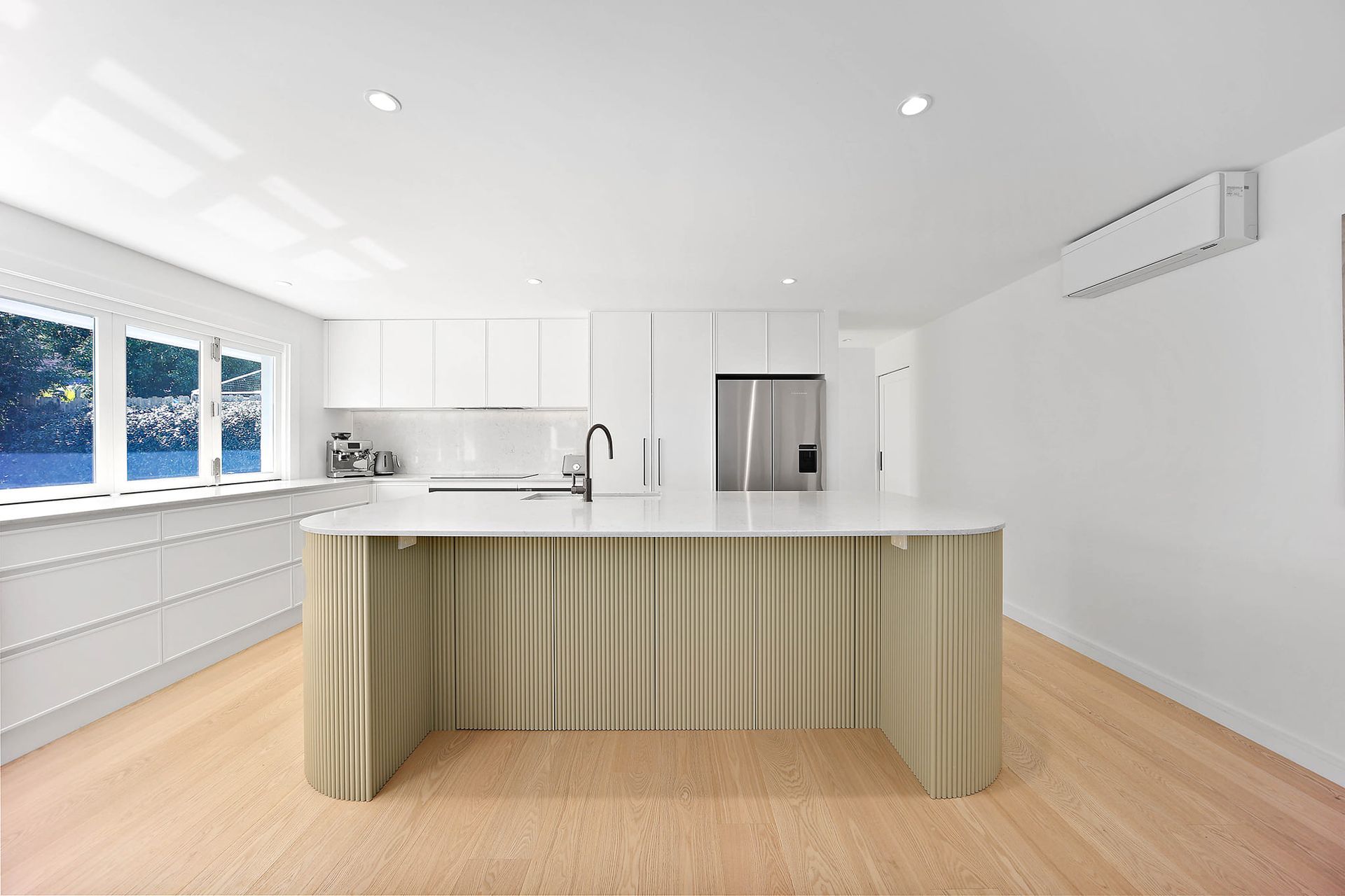 Modern white kitchen with a light wood island and flooring. A stainless steel refrigerator and large windows are visible — Above & Beyond Interiors in Wollongong, NSW