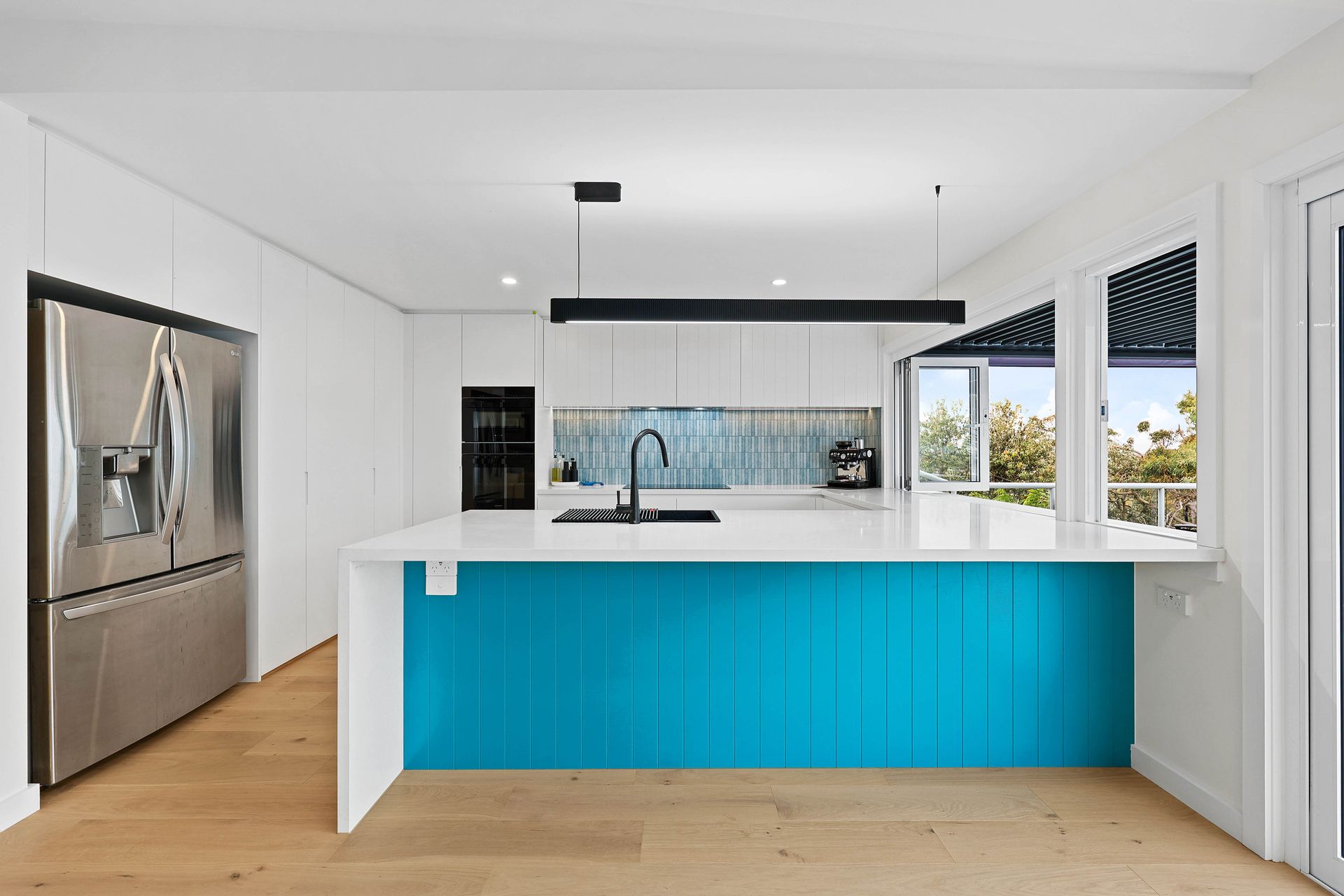A Kitchen with Turquoise Island, White Cabinets, Stainless Steel Fridge, and Large Windows — Above & Beyond Interiors in Smeaton Grange, NSW