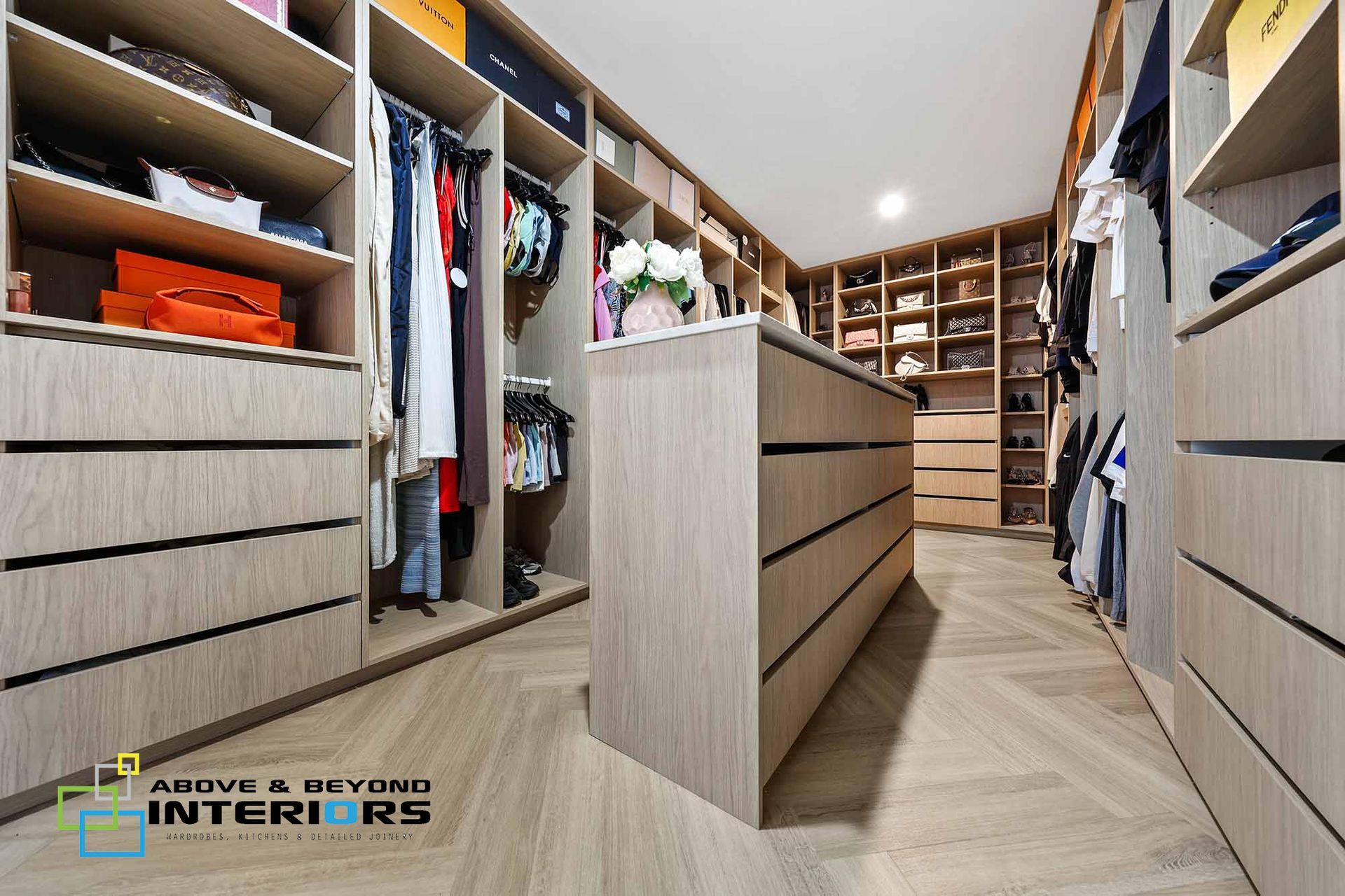 A Walk-In Closet with Wooden Shelves, Drawers, and Hanging Clothes in A Bright — Above & Beyond Interiors in Smeaton Grange, NSW