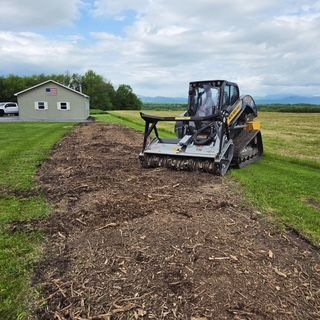 Compact track loader clearing dirt along a field beside a small house under a cloudy sky