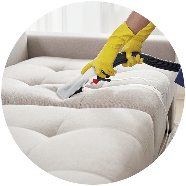A person wearing yellow gloves is cleaning a couch with a vacuum cleaner