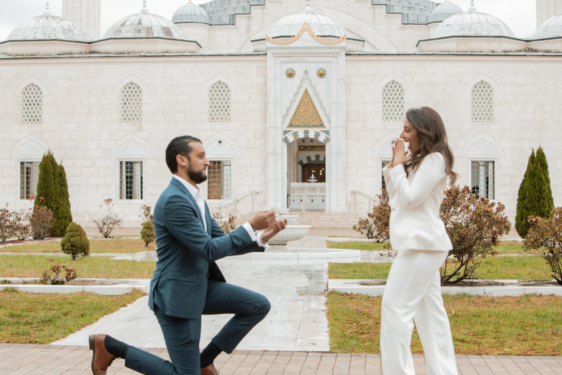 A man is kneeling down to propose to a woman in front of a mosque.