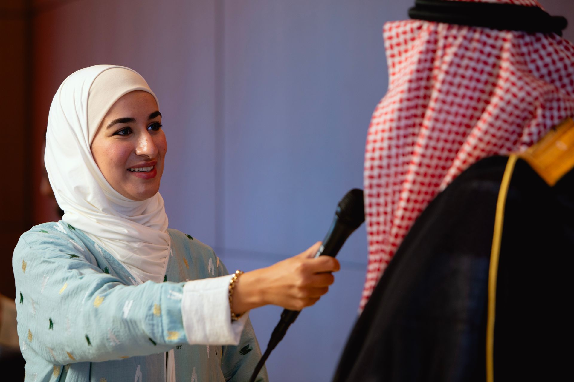 A woman in a hijab is holding a microphone in front of a man in a keffiyeh.