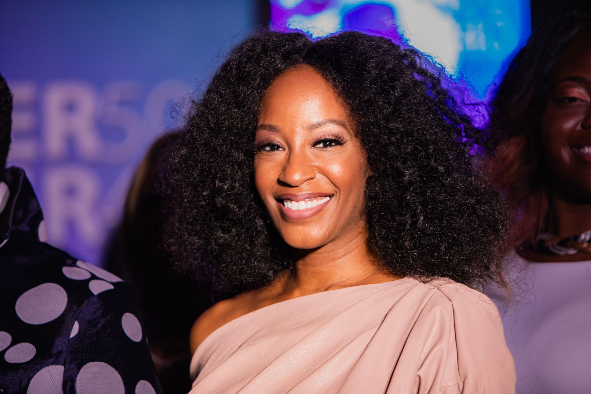 A woman with curly hair is smiling and wearing a pink dress.