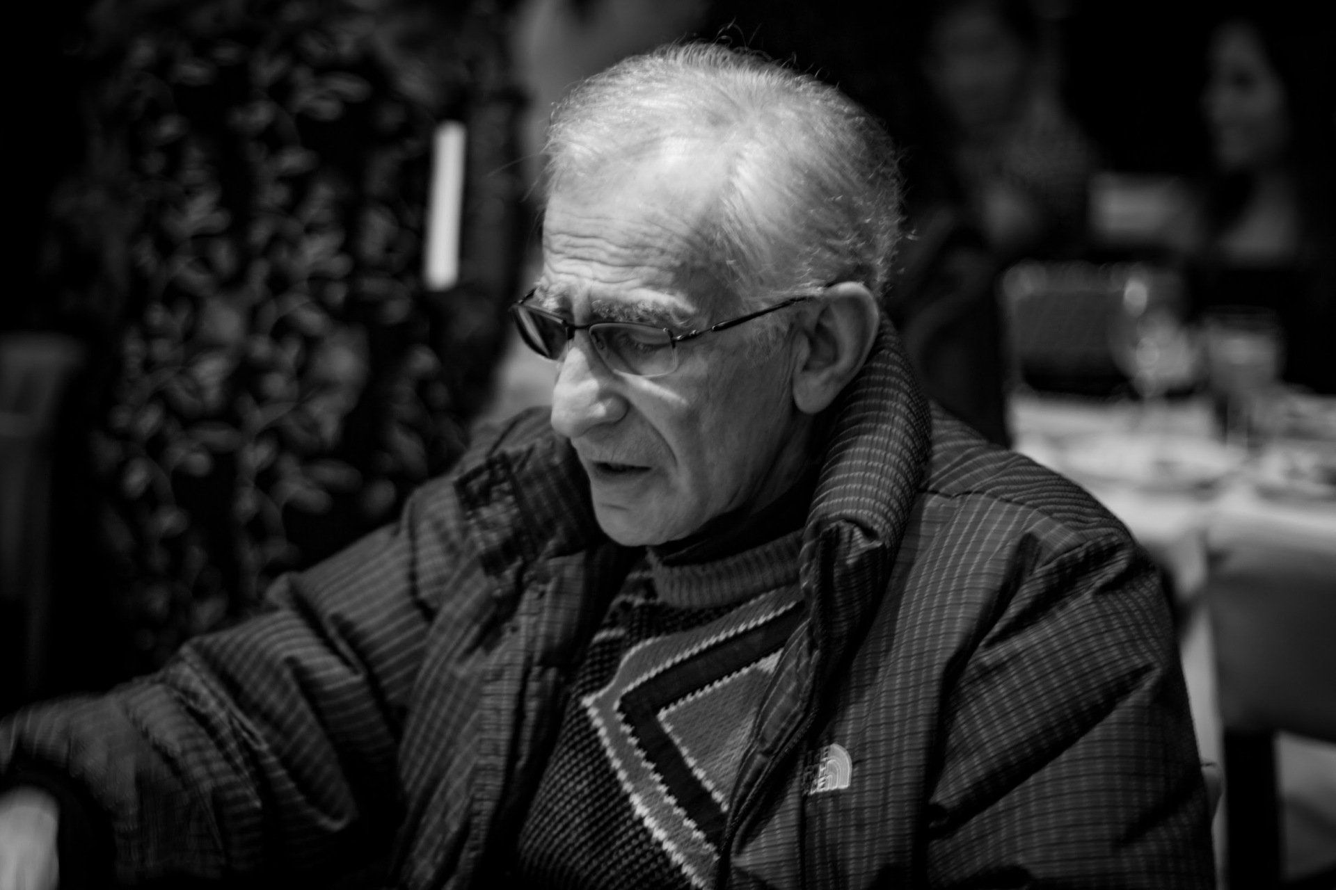 An elderly man wearing glasses is sitting at a table in a black and white photo.