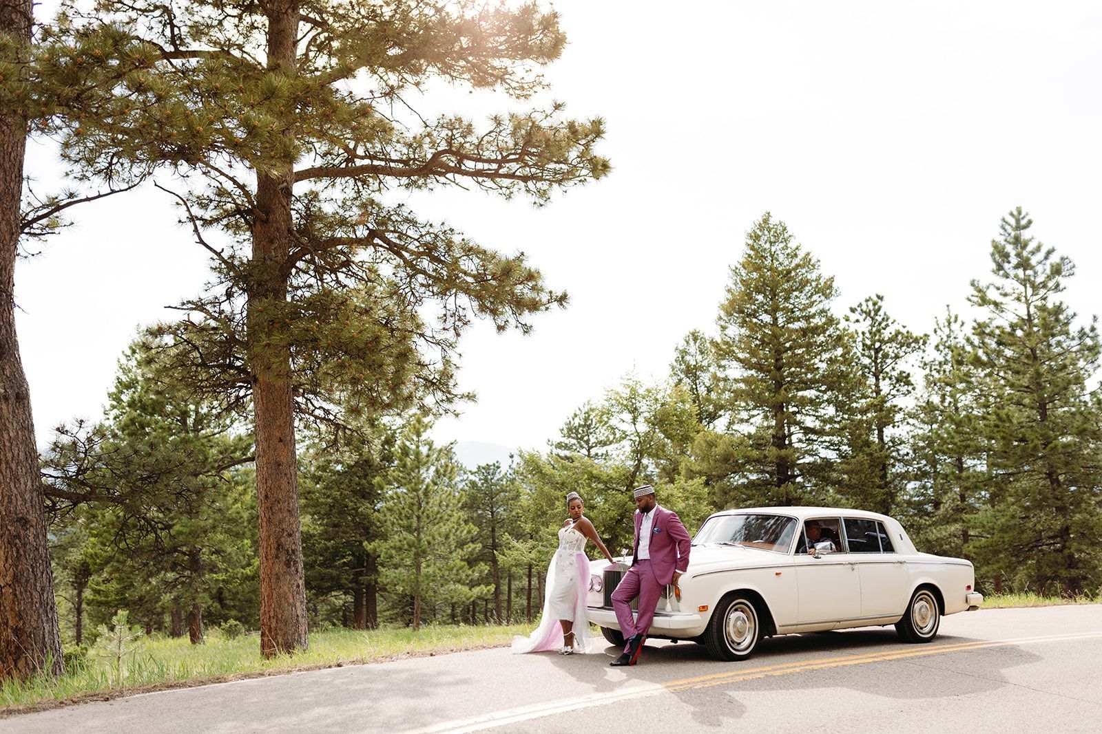 A bride and groom are standing next to a white car on the side of a road.