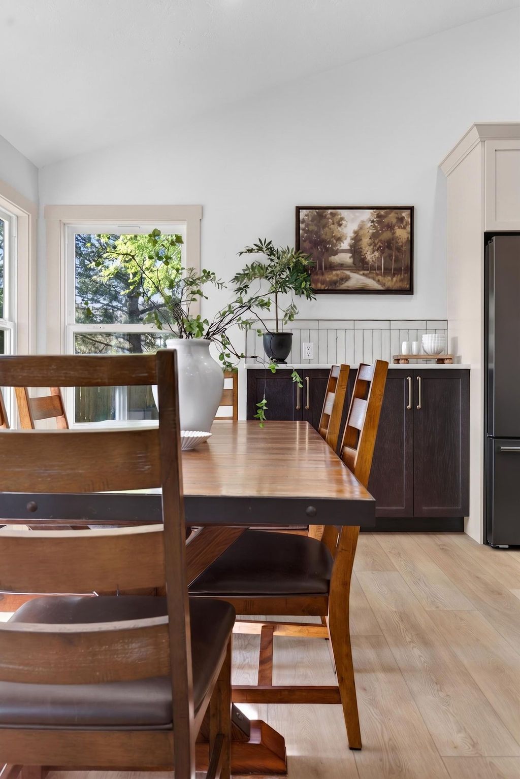 Dining room with wooden table and chairs, sideboard, wall art, and a window with plants and daylight.
